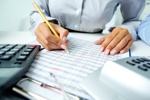 Una persona está llenando una hoja de cálculo con un lápiz en un escritorio. Sobre el escritorio también hay calculadoras y un portapapeles.