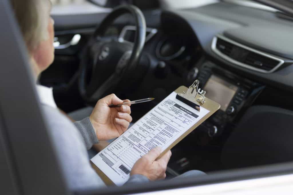 Una persona sentada en un automóvil sosteniendo un portapapeles con un formulario y un bolígrafo, probablemente completando o revisando el documento.